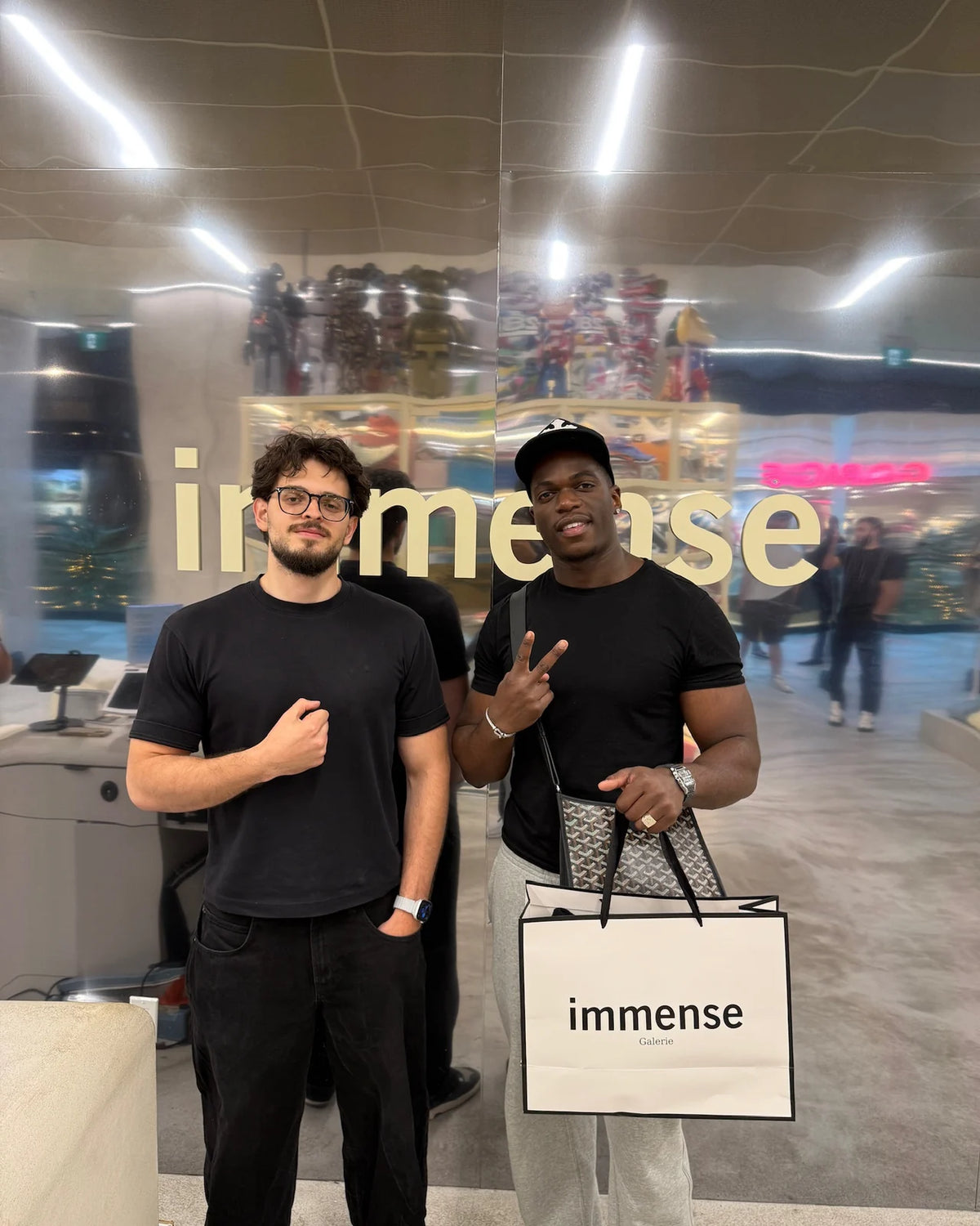 Two customers with an Immense Galerie shopping bag at the mirrored logo wall